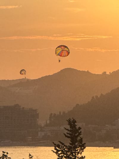 The image shows a sunset over Vrijesno, Montenegro. Two parasailers are visible in the sky, one in the foreground with a multi-colored parachute and another in the background with a smaller, similarly colored parachute. The parasailers appear to be individuals or a small group. Below them, a series of hazy, silhouetted mountains dominate the midground. In the foreground, a body of water reflects the warm, orange hues of the sunset. A large, multi-story building, possibly a hotel or apartment complex, is partially visible on the left side of the image, and a dark, silhouetted tree is on the right. The overall atmosphere is serene and picturesque, typical of a coastal sunset.