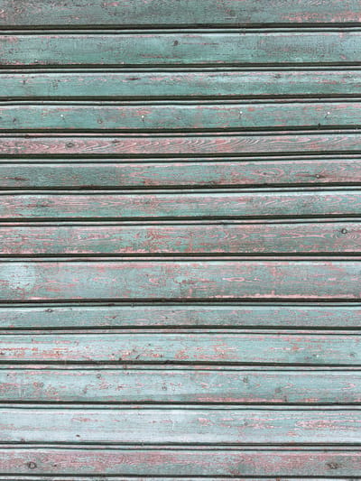 The image is a close-up, full-frame shot of a weathered wooden wall. The wall is constructed of horizontal planks with a faded, peeling teal-colored paint. The wood grain is visible beneath the paint, and there are areas where the paint has chipped away, revealing a reddish-brown underlayer. The planks are uniformly spaced, and some appear to have small nail heads or indentations. The lighting is even, suggesting an overcast day or diffused indoor lighting. There are no people or specific landmarks visible in the image, only the texture and surface of the wooden wall. The image is likely a detail shot intended for texture or material reference.