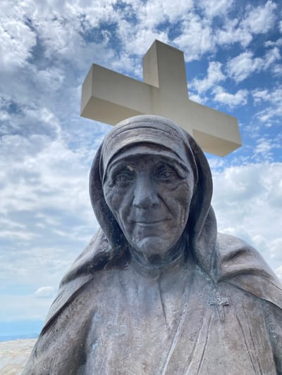 The image, captured from a low angle looking upwards, features a weathered bronze statue against a bright blue sky with scattered white clouds. The statue, positioned on the right side of the frame, depicts a woman with a serene yet solemn expression, her gaze directed slightly upwards and to the left. She wears a draped head covering and flowing garments, typical of religious habits. A small, simple cross is subtly embossed on the upper right side of her chest, integrated into the folds of her attire.
Dominating the left background is a large, minimalist white cross, characterized by its blocky, modern design. Its clean lines contrast with the texture and detail of the statue. The bright natural lighting suggests it is daytime, with fair weather. The scene creates a profound visual, evoking themes of faith and contemplation, and is located in Benkalj, Montenegro. No other text is visible in the frame.