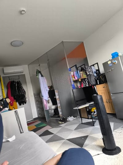 This image depicts the interior of a room, likely an apartment or dorm room, in Eschborn, Germany. The scene is filled with various objects, suggesting a lived-in space. In the foreground, a gray couch is partially visible, with a person's arm and shoulder in the lower right corner, indicating someone is present in the room. The room appears to be a studio or a multi-functional space, as it includes a kitchenette area with a silver refrigerator, and a small bathroom with a toilet and sink enclosed by a glass partition.
Various items are scattered throughout the room, including clothes hanging on a rack near a doorway, a flat-screen television on a desk, a tall black fan, and shelving units filled with personal belongings, toiletries, and food items. A brightly colored rug with geometric patterns lies on the floor, contrasting with the larger, neutral-colored rug. An air conditioning unit is mounted high on the wall. The lighting suggests it's daytime. The overall impression is one of a functional but somewhat cluttered living space. There is no explicit indication of an activity or event taking place beyond the general state of occupancy. There is no visible text in the image.