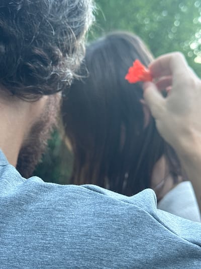 An out-of-focus image shows a person's gray t-shirt in the foreground. In the background, another person's dark hair is visible, with a blurred hand holding a small red flower near their head. The setting appears to be outdoors, possibly in a park or garden, with dappled light suggesting trees and foliage. The location is identified as Čučuci, Montenegro.
