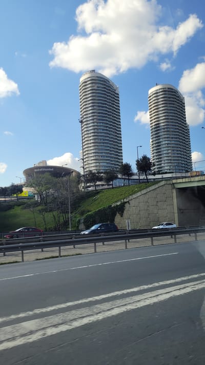 The image depicts two high-rise buildings with curved facades, suggestive of modern architecture, under a blue sky with scattered white clouds. A roadside scene is visible, including a road with multiple lanes marked by white lines. Cars are present on the road. A green embankment with vegetation and trees separates the road from the buildings. A sign reads "Mall of Istanbul". The location is Bağcılar, Turkey.