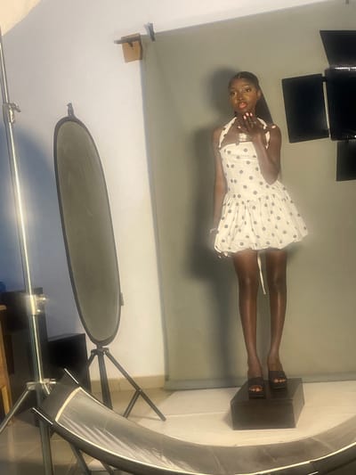 The media file depicts a young Black woman in a studio setting, posing for a photograph. She is wearing a white dress with black polka dots and black heeled sandals. The woman stands on a dark rectangular platform, with her left hand near her chest and her right hand gesturing outwards. Her expression is engaging, with her mouth slightly open and her eyes looking towards the camera.
The studio background appears to be a neutral gray backdrop. Lighting equipment is visible, including what seems to be a softbox and a reflector. The overall lighting suggests a professional photoshoot. The orientation of the image is such that the woman is standing upright, indicating it was likely taken with the camera oriented vertically. The setting appears to be an indoor studio, possibly in South Ropp, Nigeria. There is no visible text in the media file. The time of day and weather are not discernible from the image.