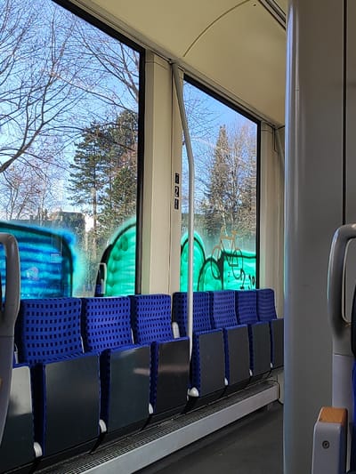 The image depicts the interior of a train or tram, likely stationary or moving slowly, given the clear external view, located in Einbeck, Germany. The perspective is from within the vehicle, looking towards the left side and outwards through two large windows.
The interior features a row of unoccupied passenger seats upholstered in dark blue fabric with a small, square grid pattern. The seatbacks are black. A light grey floor platform runs beneath the seats. A vertical cream-colored pillar with a grey support pole extends from the ceiling, dividing the windows. A small sign displaying the number "2" and a downward arrow is affixed to this pillar. The ceiling is a curved, light cream color. A partial view of another seat with a light grey handle is visible in the far right foreground.
The exterior view through the windows shows a clear blue sky and a collection of bare deciduous trees and dark green coniferous trees, suggesting a winter or early spring season. A distant building with a reddish roof and dark facade is partially visible through the left window.
A significant feature is the extensive graffiti spray-painted across the lower sections of the windows. The left window displays blue graffiti with horizontal striping. The central and right windows feature bright green amorphous shapes. Overlaid on the green graffiti on the rightmost window is a light brown outline of a bicycle. No individuals are present in the frame.