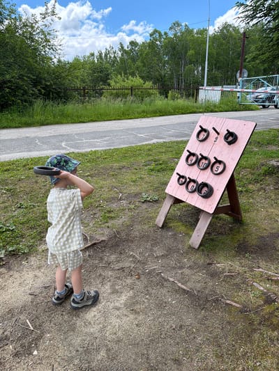 In Kul'dur, Russia, a young child stands on a gravelly and grassy area, holding a black ring in their right hand, preparing to throw it at a pink wooden board. The board, propped up on wooden legs, has pegs with rings of various sizes on them, each marked with a number from 4 to 10. The child is wearing a checkered shirt and shorts, a patterned bucket hat, and athletic shoes. Behind the child and the game, a paved path runs alongside a grassy area. Further in the background, a rusty metal fence, a white post, and a blue and white barrier gate are visible. Trees and dense foliage form the backdrop, with a bright, cloudy sky above.