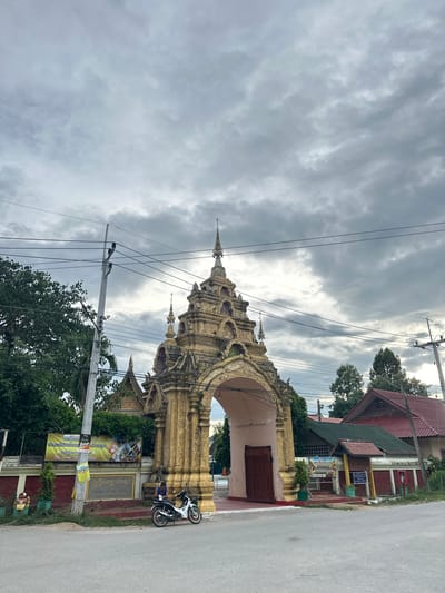The image shows a street scene in Ban Patan, Thailand. A highly ornate golden archway, likely part of a temple, dominates the left side of the frame. The arch is intricately carved with spires and decorative elements, suggesting a traditional architectural style. A paved road runs along the right side of the image.
In front of the arch, there's a man sitting on a motorcycle, which is parked on the side of the road. Behind him, a building with a red roof and a closed wooden door is visible. Several trees and electrical wires add to the urban landscape. The sky is overcast with dramatic, cloudy formations, suggesting either early morning or late afternoon, or possibly impending rain.
Several signs are displayed on the roadside and on the building. One prominent sign, with Thai script, appears to announce an event or festival. The visible text includes "พิธีงานท", "ประเพณีสรงน้ำพระ 599 นวิสาขบูชา", and "วัดลัฏฐิวนารา". This suggests a religious event related to Songkran, Vesak, or a temple anniversary. The overall atmosphere is one of a typical day in a Thai town, with elements of daily life and religious significance present.
