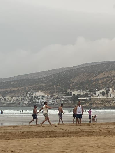 The image shows a beach scene in Taghazout, Morocco. There is a mountain and many buildings along the beach. The beach contains people and a soccer ball. Four shirtless men are playing soccer on the beach. Two people are standing in the water on the beach. One person is standing on the beach wearing a pink shirt.