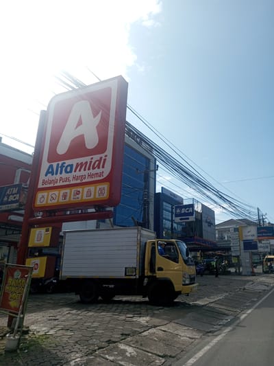 The image shows an Alfamidi convenience store with a large red and white sign. A yellow delivery truck is parked in front of the store. There's also a BCA bank sign nearby. Overhead are many powerlines, the sky is bright blue. Other signs and people are visible in the background. A sign for "Bakso Mangiae Hoci" is in the foreground. The image does not appear to be from Laos or Khe Luang and appears to be somewhere in Indonesia.