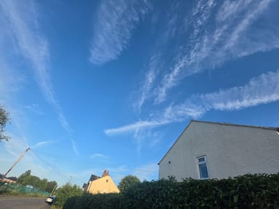 The image depicts a street scene in Shotton, United Kingdom, under a clear blue sky with wispy white clouds. In the foreground, a dense green hedge runs across the bottom of the frame. Behind the hedge, a pale yellow house with a red-tiled roof and a single window is visible on the left. To the right, a cream-colored house with a textured facade and a single window dominates the right side of the image. A street with a grey tarmac surface extends into the distance, with a silver car parked on the side. A wooden utility pole with numerous wires is prominent on the left side of the street, along with a street lamp. Trees and fences are also present in the background, suggesting a residential area. The overall impression is a quiet, daytime suburban environment.