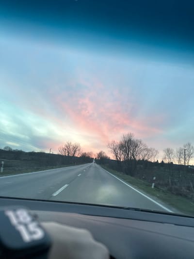 The image depicts a two-lane asphalt road extending into the distance through a rural landscape at dusk in Kovachevets, Bulgaria. The sky overhead is a deep blue, transitioning through lighter blue to a vibrant display of pink and orange stratiform clouds at the horizon, indicative of sunset. Bare deciduous trees line both sides of the road and ascend gentle, green-tinged hills. Utility poles with wires are visible along the left side of the road, and red and white roadside marker posts are present on the grassy verges. The scene is captured from the interior of a vehicle, with the dashboard occupying the bottom portion of the frame. A digital display on the dashboard prominently shows "18 15". A concentrated beam of light, consistent with a vehicle's headlight, illuminates a section of the road in the mid-ground. In the upper sky, a faint crescent moon is visible. No individuals are explicitly visible, though the perspective from within a vehicle implies a driver or occupant.