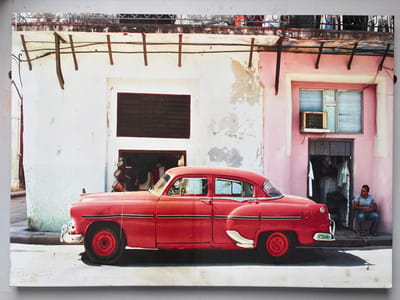 This image depicts a street scene located in the city of West Felton, United Kingdom. A bright red, vintage four-door sedan, consistent with American models from the 1950s, is parked parallel to the curb on a concrete street, oriented towards the right. The vehicle displays chrome bumpers, side trim, and red wheels.

To the left of the car, a two-story building features a white facade with extensive peeling paint and visible discoloration. An open, dark doorway below a large closed louvered window or shutter reveals an interior containing multiple conga drums and other indistinct objects.

To the right of the car, an adjacent two-story building presents a faded pink facade, also exhibiting peeling paint. Its ground-level open doorway exposes a cluttered interior with hanging garments. Above this doorway, a window with horizontal blinds is fitted with a visible window-mounted air conditioning unit.

A male individual is seated on a low stool or step just outside the pink building's doorway on the right. He wears a light-colored t-shirt and blue jeans, with his body angled towards the left.

Above the buildings, a deteriorating rooftop with a metal railing is visible, crisscrossed by numerous exposed electrical wires supported by makeshift wooden brackets. The scene is illuminated by daylight, casting shadows on the street from the car and the seated person. The overall environment suggests an aged urban setting with visible signs of wear and limited maintenance.