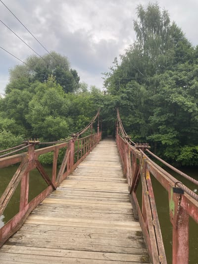 The image shows a rustic, weathered wooden suspension bridge spanning a body of water. The bridge has red metal railings and thick cables supporting its structure. The wooden planks of the bridge are worn and show signs of age. Lush green trees line the banks of the water and form a dense backdrop behind the bridge. The sky above is overcast with gray clouds. The scene appears to be set in a natural, possibly rural, environment. The location is identified as Karabanovo, Russia. There are no people visible in the image.