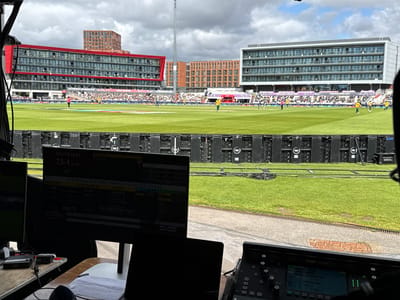 This image was taken at the Old Trafford cricket ground in Trafford, United Kingdom. In the foreground, two monitors display a cricket scoreboard with the score for Australia at "23-1". The scoreboard also shows player statistics and match information. To the right, a sound mixing console is visible. The middle ground features a lush green cricket pitch with several players in uniform, some actively engaged in the game. In the background, a large stadium filled with spectators is situated between modern buildings. The sky is partly cloudy.
