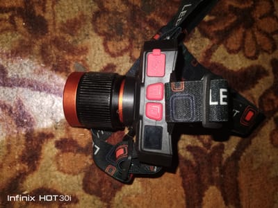 A close-up, high-angle shot shows a black and orange headlamp with a black strap that has white text and red and blue graphical elements. The headlamp is resting on a brown and tan patterned carpet. The headlamp has a bright white light shining from its front. The strap has "LED" printed on it. There are three buttons on the headlamp, two of which are red and one is black. A portion of the phone's branding, "Infinix HOT 30i", is visible in the bottom left corner of the image. The scene is indoors and it is dark, indicated by the lit headlamp. The image was taken in Lisakovsk, Kazakhstan.