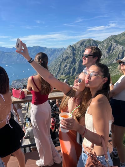 In the city of Kavač, Montenegro, two young women are taking a selfie outdoors. The woman on the left is holding up a smartphone, and both women are smiling at the camera. A man in sunglasses is visible behind them, and in the background, a scenic view of mountains and a bay can be seen. The women are holding drinks, and the atmosphere appears festive.