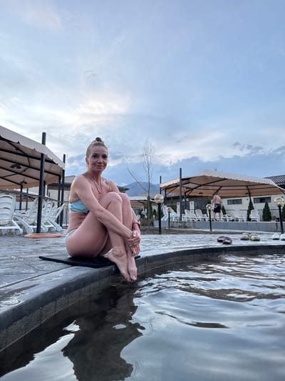 A woman is sitting on the edge of an outdoor pool in Kara-Oy, Kyrgyzstan. She is wearing a blue bikini top and holding her legs close to her body. Her hair is pulled back in a bun. In the background, there are lounge chairs under canopies, and a building. The sky is partly cloudy.
