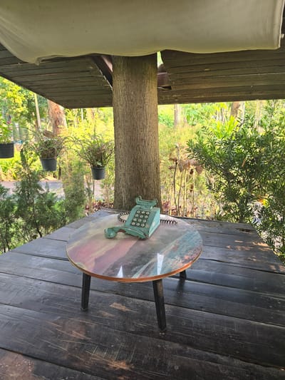 The image depicts an elevated outdoor wooden platform or deck located in Ban Phaya Kot Ana Kaeo, Thailand. The platform floor consists of dark, weathered wooden planks laid horizontally. A small, round table with a multi-colored top, featuring swirling patterns of greens, blues, pinks, and browns, stands on dark, slender legs in the foreground. Resting on the table is an antique-style, teal-colored push-button telephone; its handset is detached from the cradle and positioned beside the main unit, connected by a black coiled cord.
Above the platform, a white fabric canopy is partially visible, hanging over an underlying structure of dark wooden planks. A large, textured tree trunk rises vertically through the scene on the right, integrated into the structure. The background consists of dense, lush tropical vegetation, including various green shrubs, trees, and conifers. Several potted plants are suspended from an unseen support on the left. A narrow, paved path is faintly visible through the foliage in the mid-distance on the left side. No individuals are present in the scene.