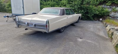 A cream-colored, vintage Cadillac Eldorado is parked on an asphalt surface. The car is viewed from the rear-right quarter. The license plate reads "C-1211". A white trailer with a blue hitch mechanism is attached to the rear of the car and extends to the left of the frame. The background consists of dense green foliage and a rocky embankment. The scene is situated in a suburban or rural setting, likely on the outskirts of the city of Ørsta, Norway. The lighting suggests it is daytime, with shadows cast on the asphalt.