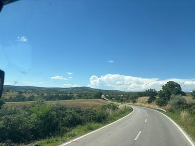 The image captures a curving asphalt road passing through a rural landscape under a bright blue sky with scattered white clouds. The road is bordered by dry grass and vegetation on the right, and a grassy verge with some bushes on the left. Beyond the road, rolling hills dotted with trees and fields stretch into the distance. The scene is devoid of any people or discernible structures other than the road and guardrails. The presence of dried grasses suggests a warm weather season. This road is identified as being in the vicinity of Mladinovo, Bulgaria.