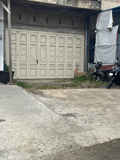 This is a medium shot, eye-level, natural light, outdoor photograph of a store front in the city of Lampisang, Indonesia. The store front features large, off-white, double doors with decorative panels. Above the doors is a concrete overhang with small, rectangular ventilation slots. To the right of the store front, a blue metal gate is visible, with white tarpaulins hanging behind it. In front of the store, two black motorcycles are parked. The ground is a mix of cracked concrete and gravel, with patches of green grass growing in some areas. A black plastic planter is located to the left of the store entrance. The overall scene depicts a typical urban streetscape with commercial activity.