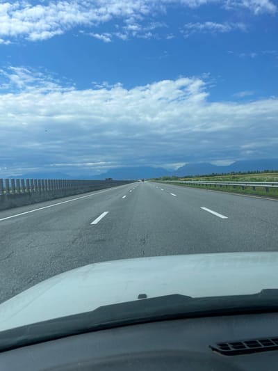 A first-person perspective from a vehicle driving on a highway. The highway has multiple lanes, marked by white lines, and is bordered by a concrete barrier on the left and a guardrail on the right. Beyond the barrier, a row of cylindrical posts is visible. In the distance, mountains form a hazy silhouette against a sky filled with both blue patches and dark, cloudy formations. A single bird can be seen flying in the sky on the left side of the image. The hood of the vehicle is visible in the foreground, obscuring the lower portion of the scene. There is no specific indication or identifiable feature within the image that places the location in Alkhanchurt, Russia.