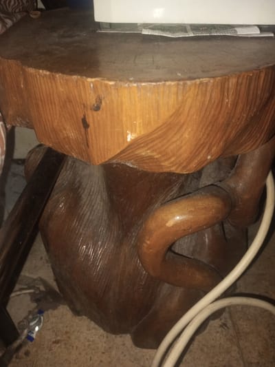 A close-up image shows a round, wooden side table with a flat top surface. The table is carved with a rustic, naturalistic design. A white electrical cord is draped near the base. The location of the image is Yin Gat Kone, Myanmar.