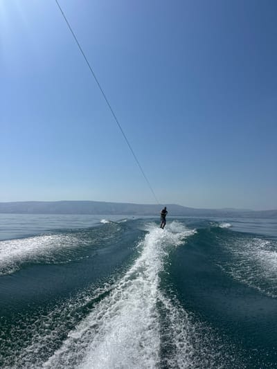 The image depicts a male individual wakeboarding on the Sea of Galilee, in the vicinity of Tiberias, Israel, under a clear blue sky. The upper half of the frame is dominated by a cloudless, light blue sky. A thin, dark tow rope extends diagonally from the upper left towards the center, attaching to the wakeboarder. The lower half of the frame shows the dark blue-green water of the lake, with significant white foam and turbulent wake generated by a moving boat, from which the image is presumably captured, and the wakeboarder.
The wakeboarder is positioned towards the middle right of the frame, standing upright on a dark wakeboard, which is partially obscured by spray. The individual wears a dark short-sleeved top and dark shorts. The wakeboarder's body is slightly angled, suggesting active balance and control. Behind the wakeboarder, a prominent, foamy white wake extends towards the bottom of the frame, indicating rapid movement. In the distant background, a low-lying range of hazy hills or mountains is visible along the horizon line, separating the water from the sky.