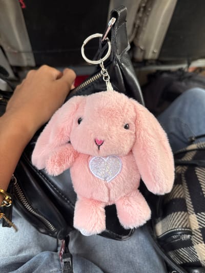 A fluffy pink bunny keychain is attached to a black leather bag, with a silver keychain ring and chain visible. The bunny has a pink heart nose and a glittery heart design on its chest. A hand with a bracelet is on the left side of the image, resting on the bag. The background shows part of a person's leg in blue jeans and a black and cream plaid blanket. The scene appears to be inside a vehicle, possibly a bus or van, given the visible metal and fabric textures in the background which are consistent with public transportation seating. The overall lighting suggests an indoor environment. There is no specific indication of the Santa Cruz, Philippines location within the image.