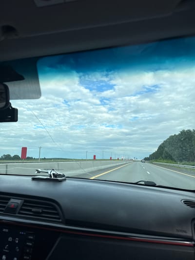 The image shows a view from the interior of a car, looking out through the windshield. The car is on a highway, with multiple lanes of traffic visible. The sky is cloudy, with patches of blue sky showing through. There are trees and fields on either side of the highway. A small, silver helicopter-shaped ornament is visible on the dashboard of the car, to the right of the steering wheel. A dashcam is mounted on the windshield, to the left of the rearview mirror. The time "19:05" is visible on the car's dashboard display. The location is stated to be the city of Можга, Russia.