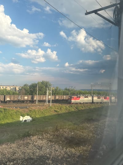 The image shows a train passing through a rural area with a town or city in the background. The train consists of a locomotive and several freight cars. The locomotive is red and white with the Russian Railways logo. The freight cars are dark brown. In the background, there are several multi-story buildings and trees. The sky is blue with white clouds. The foreground shows a grassy field with some dry vegetation. There is a concrete structure with a circular opening in the grass. The image is taken from a window, as evidenced by the slight blur and reflection on the right side of the frame. The scene appears to be in Syzran, Russia, given the distinctive architecture of the buildings and the style of the train.