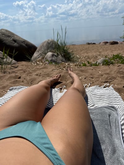 The image displays a close-up, low-angle view of a person relaxing on a sandy shore, seen from the waist down. The person's legs and feet are prominent in the foreground, with their right leg partially obscured. They are wearing a light blue or teal bikini bottom. Beneath them are two towels: one with grey and white stripes, and another dark grey, textured towel, both casting shadows suggesting bright sunlight.
The setting appears to be a beach or lakeshore. The ground is sandy, interspersed with small rocks and patches of green grass, particularly on the left side of the frame. In the background, beyond the sandy stretch, a calm body of water is visible, meeting a clear sky dotted with white cumulus clouds. The bright lighting indicates a sunny daytime scene. The overall activity depicted is one of leisure and relaxation, likely sunbathing. There is no visible text in the image.
