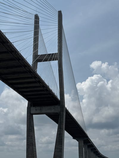 The image shows a low-angle perspective of the Sidney Lanier Bridge in Brunswick, Georgia, USA. The bridge is a cable-stayed bridge with two tall concrete pylons. The road deck of the bridge is visible in the foreground, extending from the left side of the image towards the right. Numerous white cables run from the tops of the pylons to the road deck, creating a striking geometric pattern against the cloudy sky. The pylons are massive concrete structures with a stepped design. The sky is partly cloudy, with a mix of bright white cumulus clouds and darker, more diffused clouds. The overall impression is one of immense scale and engineering prowess.