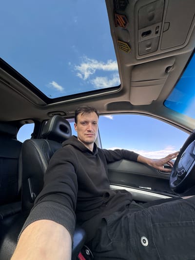 A man is seated in the driver's seat of a car, positioned in what appears to be the city of Abrykosivka, Ukraine. The car has a sunroof open, revealing a bright blue sky with scattered white clouds. The man, wearing a black hooded sweatshirt and dark pants, is looking directly at the camera. His left arm is resting on the car door, and his right hand is on the steering wheel. The interior of the car is visible, including black leather seats and the dashboard. The overall scene suggests a daytime setting, likely during fair weather.
