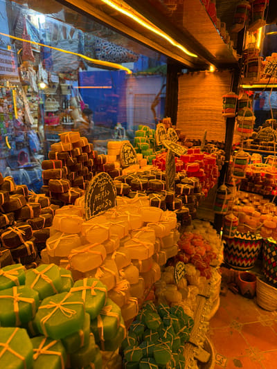 The image presents the brightly lit interior of a market stall or shop in Chefchaouen, Morocco, prominently featuring a diverse display of artisanal soaps. The foreground is filled with numerous stacks of rectangular soap bars, individually wrapped with natural-colored string or raffia. These soaps exhibit a wide spectrum of colors, including vibrant green, translucent yellow/amber, dark brown, red/pink, and lighter off-white/cream. Small, dark, irregular-shaped signs with white chalk-like script are interspersed among the stacks, identifying specific varieties like "Sabon d'Herbs Louisa" (Lemon Verbena soap) and "Sabon d'Fleur d'Oranger" (Orange Blossom soap).
Warm, orange-toned artificial lighting, including a prominent linear LED strip along the upper display edge, illuminates the scene. To the right, additional soap displays are visible, along with colorful, woven cylindrical containers holding more soaps and decorative woven baskets with geometric patterns on a tiled floor.
Through the glass storefront, reflections of the shop's interior overlay an exterior view of Chefchaouen's distinctive blue-painted buildings, characteristic of the city's medina. A faint, blurred view of another shop or stall across the street can be seen, displaying hanging textiles and bags, one with the word "SOLDES" (Sales) visible. A vague, indistinct human form is barely discernible in the background, either as a reflection or through the window. No clear actions or interactions are depicted within the immediate shop space.