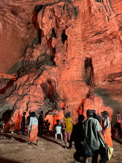 The image depicts a dimly lit, cavernous space with rough, reddish-orange rock formations that resemble a cave. The cave walls and ceiling are textured with natural geological features, and the space is illuminated by artificial red lighting, casting dramatic shadows. In the foreground and midground, several individuals are present, appearing to be visitors or participants in an event.
On the left side, a person in a blue shirt and dark pants stands facing away from the camera. Next to them, another person wears a light gray top and an orange lower garment. Further into the midground, a group of people are gathered, some standing and some seated. One person in the center is wearing a bright yellow and black patterned garment with red lower garments. Another individual sits on a white chair.
In the background, a tall, wooden ladder is propped against the cave wall, leading up to a dark opening. A person in an orange garment is visible near the base of the ladder, seated on a rock formation. The cave floor appears to be dirt or gravel. Scattered around the space are some objects that might be religious or decorative items, including what appear to be trident-like structures standing upright in the ground.
The overall scene suggests a visit to a significant natural landmark or a place of cultural or religious importance, possibly a cave shrine or a tourist attraction within the Gollagutta region of India. The presence of multiple people and the lighting indicate some form of organized gathering or tour.
