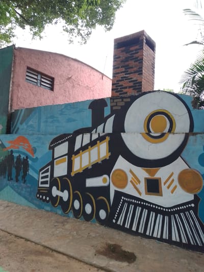 The image depicts a mural painted on a wall in Villa Hayes, Paraguay. The mural features a stylized depiction of a vintage steam train, painted in black, white, and yellow. To the left of the train, silhouettes of soldiers march towards an orange sunrise or sunset. Above the mural, there is a pink building with a boarded-up window and a brick chimney. The ground in front of the wall is paved. Some foliage is visible in the top left corner.