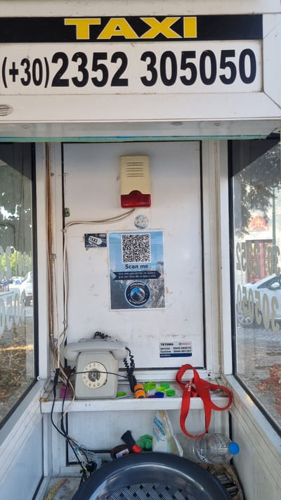 The image shows the interior of a small, enclosed booth, likely a taxi stand or a telephone booth, in Litochoro, Greece. The booth has glass panels on the sides, offering a view of the exterior, which appears to be a street with trees and buildings.
Dominating the top of the booth is a large sign with "TAXI" written in yellow on a black background, followed by a phone number "+30 2352 305050" in black text on a white background.
Inside the booth, a rotary telephone is mounted on a shelf. To the right of the telephone is a QR code poster with the text "Scan me" and Greek writing below it, along with a logo for "LITOCHORO". Above the QR code is a cream-colored alarm or siren device with a red light.
Various objects are scattered on the shelves and floor of the booth, including a red strap, plastic bottle caps, a small brush, a bag labeled "SUMMIT GREENWASH", and a plastic water bottle. Wires and cables are visible, connecting to the telephone and the alarm device.
The overall condition of the booth suggests it is functional but somewhat aged and cluttered. The environment outside the booth appears to be an urban or semi-urban setting.