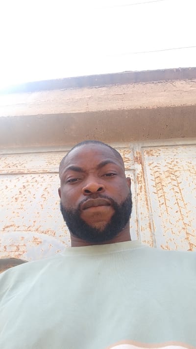 An adult male is captured in a close-up, low-angle portrait, purportedly located in Dandagoro, Nigeria. He has dark skin, short dark hair, and a full, dark beard. He wears a plain, light pale green crew-neck shirt. His gaze is directed forward, toward the camera. The background consists of a vertical surface, possibly a metal gate or wall, exhibiting significant signs of wear, characterized by peeling light-colored paint and extensive reddish-brown rust or discoloration. Above this surface, a rough-textured, light brown concrete beam spans horizontally. The uppermost portion of the frame shows a bright, overexposed white sky, suggesting strong overhead daylight, with faint, thin horizontal lines, likely overhead wires, visible against it.