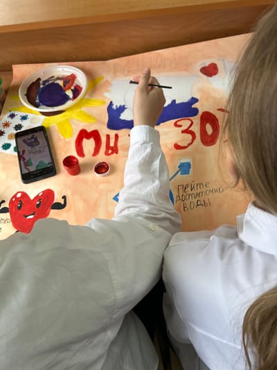 Two individuals, observed from an overhead perspective, are seated at a table in the city of Raduga, Russia, engaged in painting a large poster. The individual on the left, partially visible, is wearing a white long-sleeved shirt. The individual on the right, also in a white shirt, has long brown hair and is actively holding a paintbrush, applying white paint to a blue-colored section of the poster.

The poster, laid flat on a light-colored surface, features various painted elements. These include a large yellow sun-like image, several red, blue, and white painted shapes, the Cyrillic letters "MЫ" (MY) in red, and the number "30" also in red. To the lower left, a stylized red heart character with flexed arm muscles is depicted. Below the "30," a drawing of a blue glass of water accompanies the handwritten Cyrillic text "ПЕЙТЕ ДОСТАТОЧНО ВОДЫ" (Drink enough water). Small, colorful, abstract shapes resembling microbes are present near the sun figure.

Adjacent to the poster, a white plastic container holds various paint colors, including shades of blue, red, brown, and white. Two small open pots of red paint are positioned nearby. A black smartphone with a colorful cartoon image and Cyrillic text on its screen is placed to the left of the painting area. A light brown wooden surface is visible in the upper background.