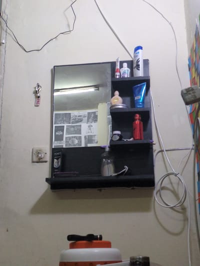 The image shows a wall-mounted shelf unit with a mirror, located in Abbottabad, Pakistan. The shelf unit is dark in color and has two shelves. On the top shelf, there are several toiletries including a bottle of shampoo, a small white bottle with a blue label, a tube of cream, and a pink bottle. On the lower shelf, there is a blue tube of lotion, a red metallic bottle, a dark blue jar, and a clear pump bottle. A pink brush is visible on the shelf. The mirror reflects a fluorescent light and a collage of black and white photographs. Below the shelf unit, on the wall, is a light switch. Further down, a white water cooler with an orange and white lid and a black handle is visible, with a metal object on its side. The wall is painted a light color and is partially covered by electrical wires. A key with a tag is hanging on the wall to the left of the shelf.