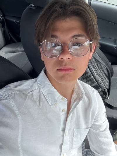 A young man with brown hair and light facial hair wears silver-rimmed glasses. He is in a car, reclined in a seat. He is wearing a white button-down shirt, partially unbuttoned at the top. A car seat and a black and grey patterned bag are visible in the background. The image is taken from a slightly high angle, looking down at the subject.