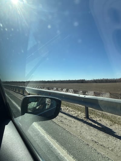 The image is taken from inside a moving vehicle. The scene shows a roadside view featuring a metal guardrail, a field, and a clear blue sky. The sun's glare is visible through the windshield. The vehicle's side mirror reflects the driver. The location is not identifiable as within the city of Обухово, Russia from the imagery.