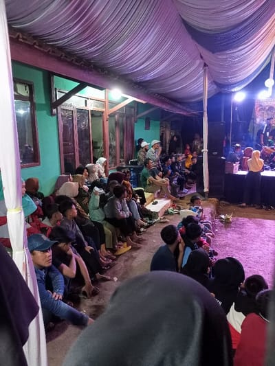 This media file depicts a nighttime community gathering or performance in Kecamatan Cimaung, Indonesia. A diverse group of people, including men, women, and children, are seated on the ground, steps, and benches under a temporary canopy with draped white and purple fabric.

The setting is an outdoor or semi-outdoor space, with a distinct turquoise-walled building featuring wooden windows and doors in the background. Artificial lighting illuminates the scene, with bright stage lights casting a purple hue on the performance area to the right, and overhead white lights providing general illumination.

The attendees are focused on a stage where performers are visible. One performer, possibly a woman in a yellow or cream hijab and dark attire, is seen from behind near musical instruments, including drums. Large speakers flank the stage, and a banner with partially visible text is displayed as a backdrop.

Most audience members are seated casually, many having removed their footwear. The atmosphere appears relaxed and attentive, as the community observes the event. A blue water bottle is visible among the seated individuals. The foreground is partially obscured by the head of a person seated close to the camera. The ground where the audience sits consists of both concrete and bare earth.