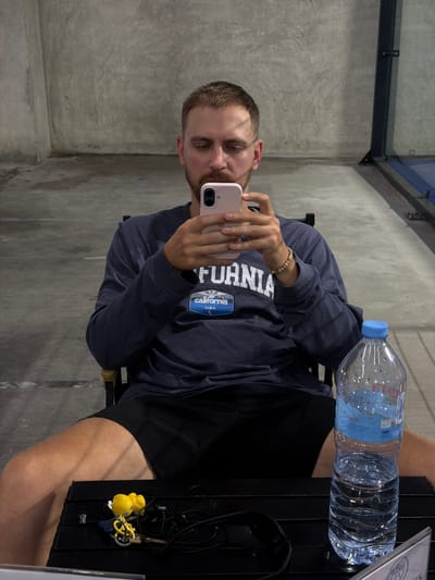 A man sits in a director's chair, looking at his pink smartphone, in front of a textured gray concrete wall. He wears a dark blue long-sleeved shirt with a "California USA" graphic and black shorts. On a black table in front of him, there's a clear plastic bottle of water with a blue cap and a label depicting mountains, along with a set of keys and earbuds. The floor appears to be concrete, and a glass partition is visible in the background on the right. The scene is likely at a sports facility, given the glass partition that could be part of a court. The provided location of Ban Buak Khrok North, Thailand, suggests this is an indoor or covered outdoor area, possibly an air-conditioned space given the man's attire.