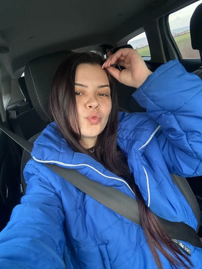 A female subject is depicted seated in the front passenger seat of a vehicle in Padarevo, Bulgaria. She is wearing a royal blue quilted jacket with white trim and is secured by a grey seatbelt, which displays an 'AIRBAG' label near her left shoulder. Her long, dark hair is parted on the left and falls over her shoulders. Her face, characterized by fair skin and visible freckles across the nose and cheeks, is directed towards the viewer, with her lips pursed in a kissing expression. Her left hand is raised, with light-colored fingernails touching her temple/hairline. The vehicle's interior is dark, and the external environment, viewed through the rear passenger window, consists of green agricultural fields extending towards distant low hills under an overcast sky.