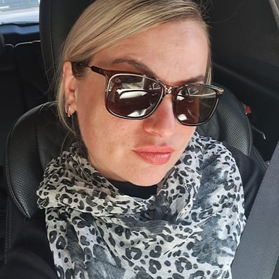 A middle-aged, fair-skinned woman is reclining in a black leather car seat. She is wearing brown-framed sunglasses perched on her nose, and a black and white leopard-print scarf is draped around her neck and chest. She has blonde hair and a small gold hoop earring in her left ear. The interior of the car is visible in the background, including a seatbelt. The location in Elektrougli, Russia, cannot be confirmed from the image.