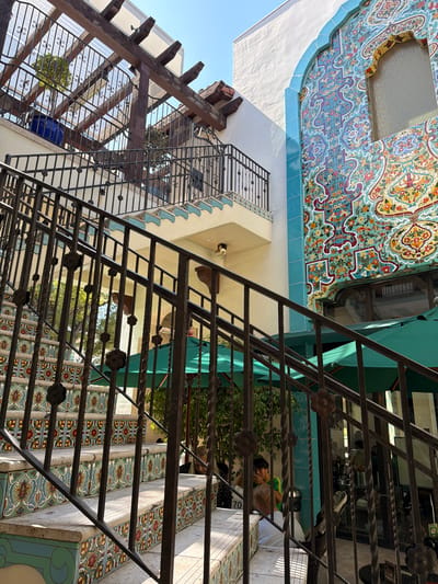This image shows an outdoor staircase with ornate tilework on the risers and edges, located in Pasadena, California, United States of America. The staircase has a black wrought iron railing. Above the staircase, there is a wooden pergola structure with a translucent covering and a balcony with similar tiling and railing. To the right of the staircase, a white building is adorned with vibrant, intricately patterned mosaic tiles, featuring a large arched window. Green outdoor umbrellas are visible beneath the staircase, suggesting an outdoor seating area. Several people are present, with some walking up or down the stairs and others visible in the background, possibly seated at tables. The lighting suggests it is daytime, with bright sunlight casting shadows.