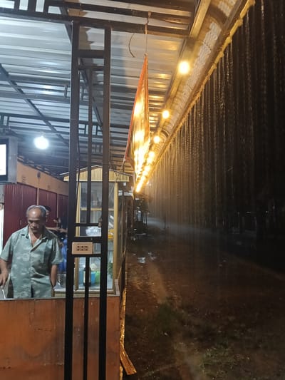 This image depicts an outdoor scene at night in Blangkutacoh, Indonesia, during a heavy rainstorm. A man, appearing to be middle-aged with a bald head and wearing a light green patterned short-sleeved shirt, stands under a covered structure. He is looking down and appears to be working or preparing something at a counter. Behind him, a stall or food cart is visible with some containers and a metal pitcher.
The setting is an outdoor marketplace or street with various stalls. A corrugated metal roof is supported by dark metal beams, providing shelter. String lights, a row of yellow-toned bulbs, are illuminated along the edge of the structure, casting a warm glow. These lights are partially obscured by the heavy rainfall, which is depicted as streaks of water falling from the roof and creating a misty effect. The ground appears to be wet and muddy, with puddles reflecting the faint light.
To the right of the frame, the rain is the dominant feature, creating a curtain of water that obscures the background and emphasizes the dark, rainy night. The contrast between the illuminated stalls and the dark, rainy atmosphere suggests a somewhat subdued or perhaps cozy ambiance, despite the downpour. The man's expression is neutral, and the overall mood is one of ongoing activity within a challenging weather condition. No visible text is present in the image.