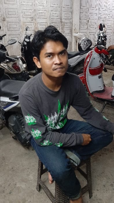 An adult male, approximately 20-30 years old, with dark, short hair and a light mustache, is seated on a brown, woven stool, facing the camera with a neutral expression. He is wearing a dark grey long-sleeved t-shirt with green and white stylized leaf graphics on the chest and sleeves, and blue jeans rolled up at the ankles, revealing bare feet resting on a concrete floor. The scene is located within a garage or sheltered area in RawangItiek, Indonesia. In the background, multiple motorcycles are parked: a light-colored scooter is partially visible to the left, and a red and white scooter (possibly a Honda Scoopy) with a white full-face helmet resting on its seat is to the right. A dark-colored motorcycle is also visible behind them. The back wall is light-colored, possibly white, featuring a repeating embossed rectangular pattern and appears weathered. A white ceramic sink is affixed to the wall, visible above the red and white scooter.