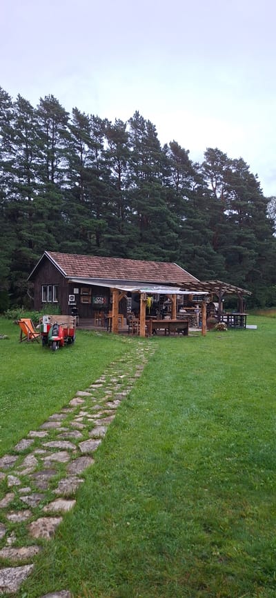 In Orissaare, Estonia, a rustic wooden building, possibly a restaurant, is situated in a grassy field. The building has a tiled roof and a covered outdoor seating area furnished with wooden tables and chairs. In front of the building, a stone pathway leads through the lawn. To the left of the pathway, an orange lounge chair is placed next to a vintage red and green three-wheeled vehicle with a wooden cargo box. A dense forest of tall pine trees forms the backdrop of the scene, and the sky is overcast.